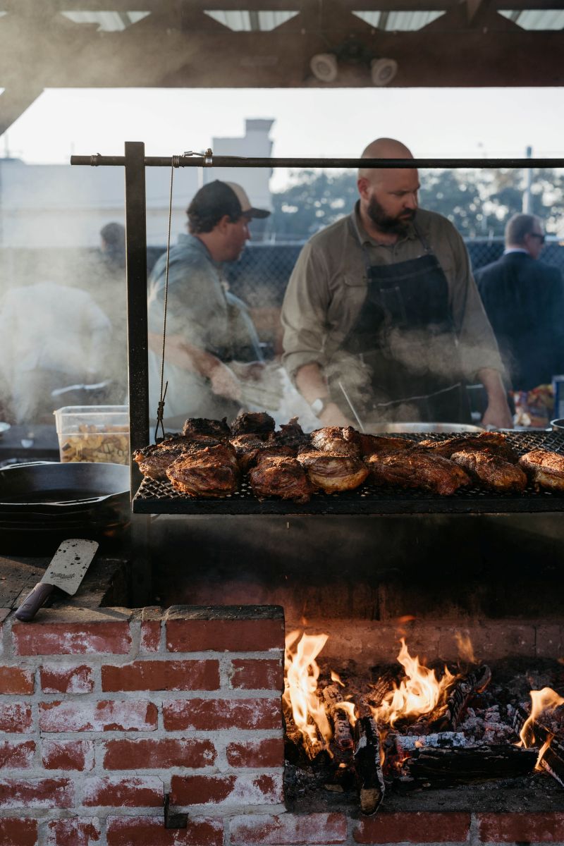 chefs at an outdoor grill cooking meat and underneath is a firepit 