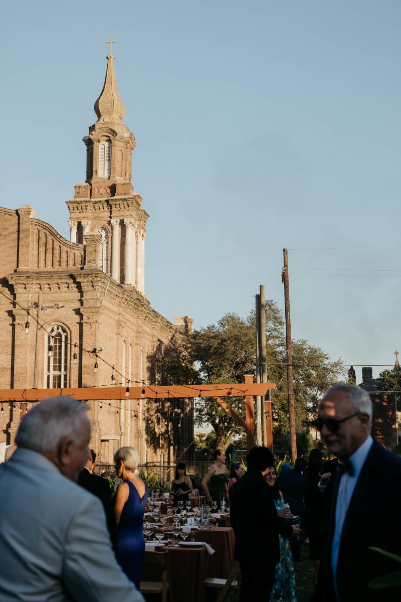 guests mingling at an outdoor wedding there is a church behind them and above them there are string lights 