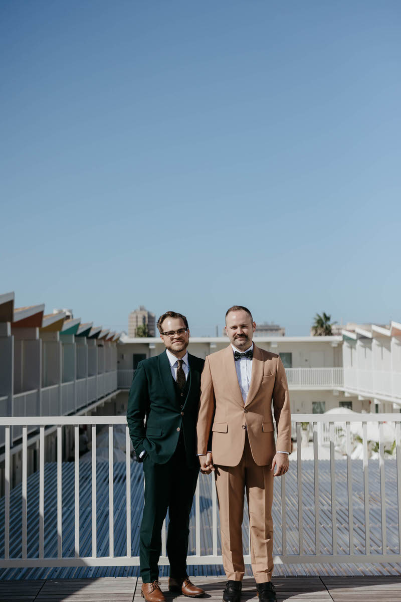 A newlywed couple in suits holding hands and standing on a hotel balcony looking over a pool 