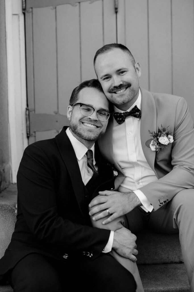 Two grooms sitting on stairs leaning into each other and smiling 