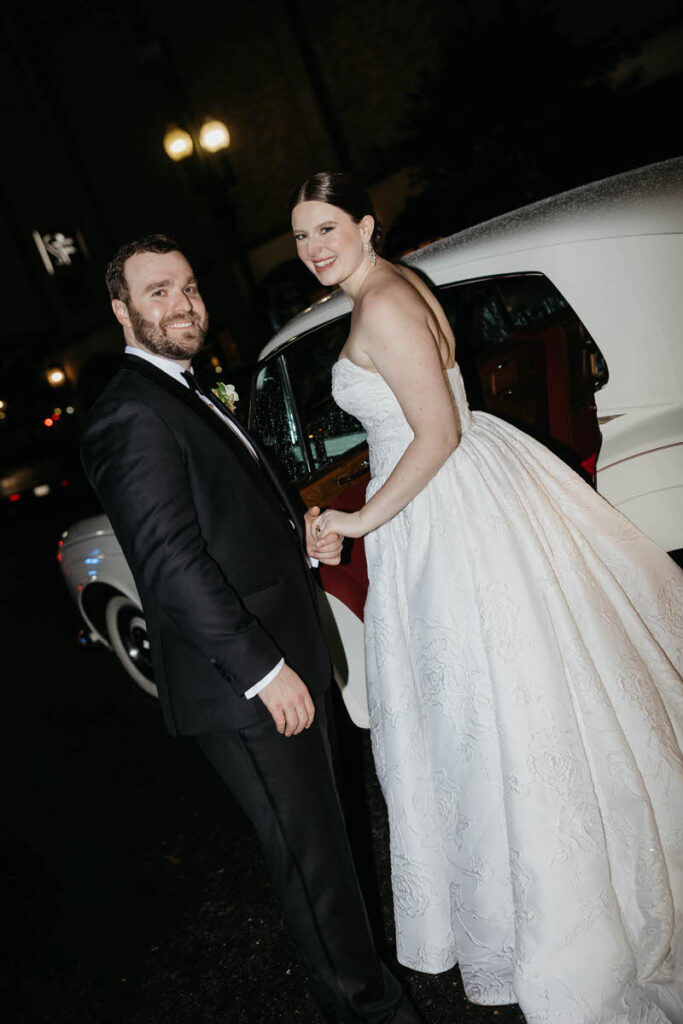 A newlywed couple holding hands and smiling as they step in to a car.