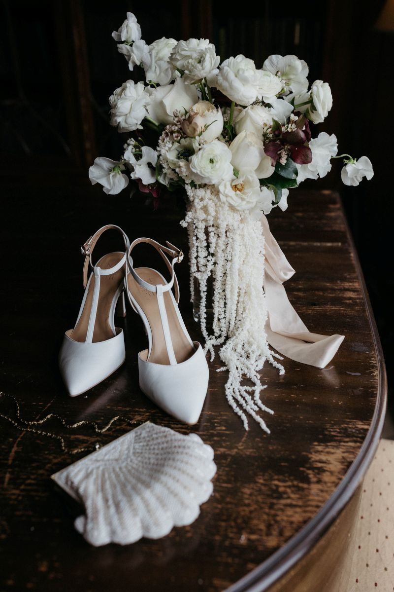 wedding details including an elegant bouquet of flowers and white heels resting on a table along with a clutch purse