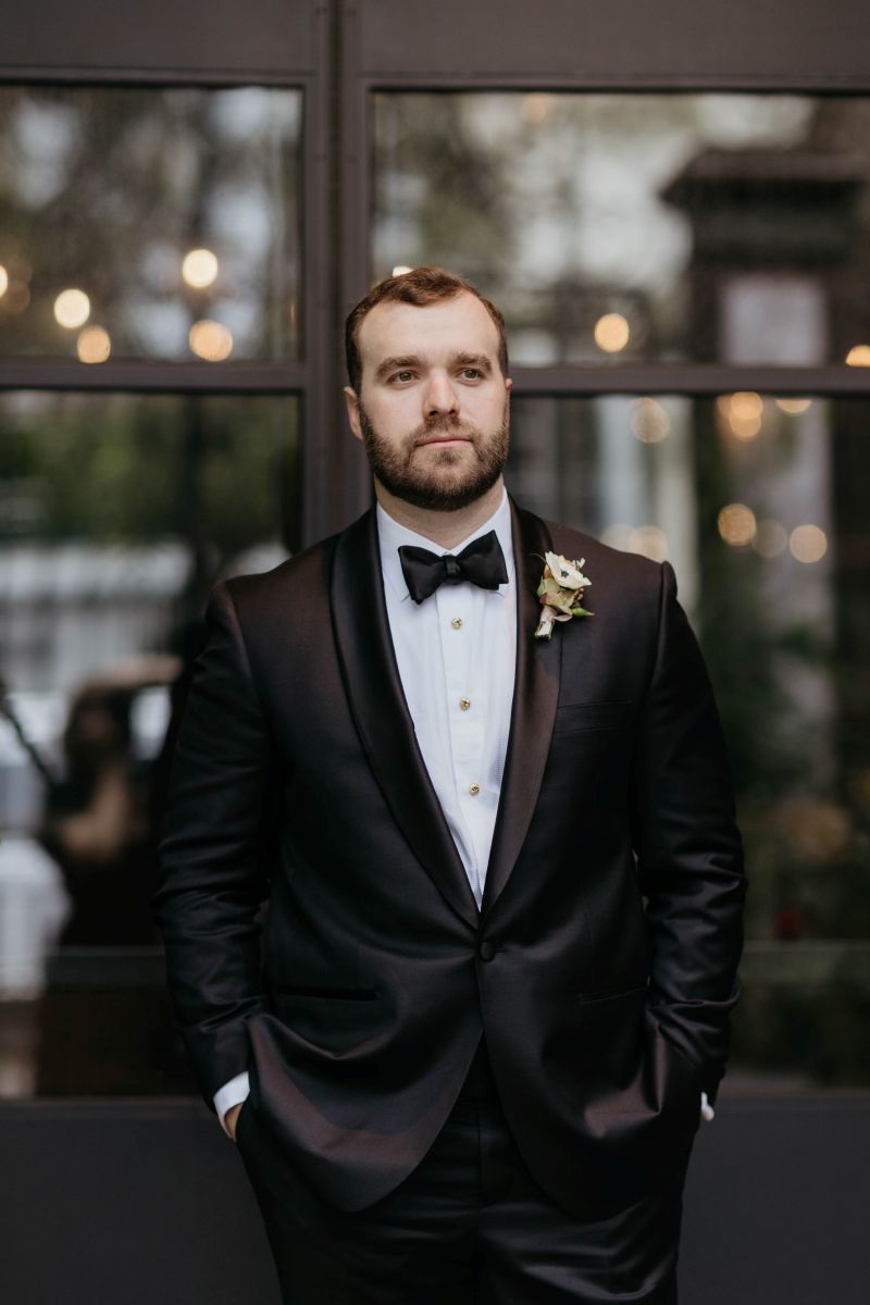 a man posing in front of a window with his hands in his pockets he is wearing a black suit