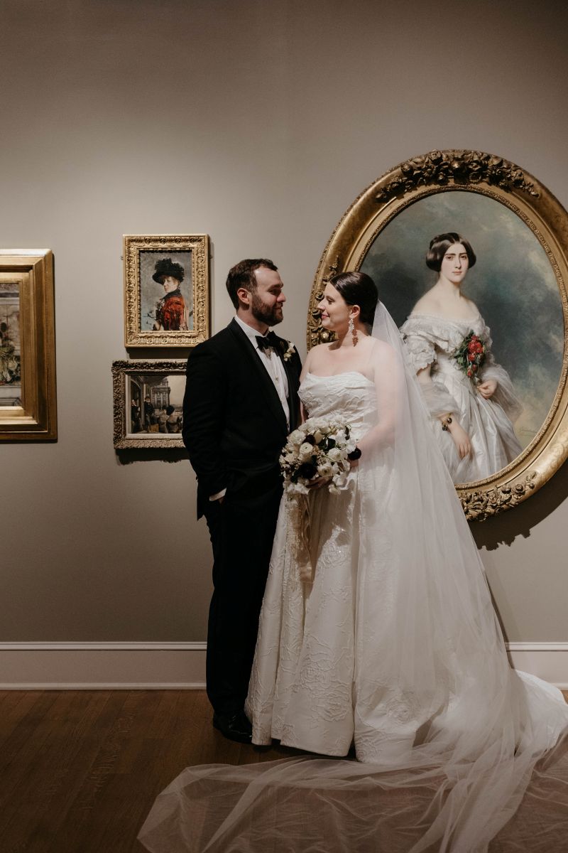 a couple posing in front of art in the new orleans museum of art the woman is wearing a white wedding dress and veil and holding flowers the man is wearing a black suit