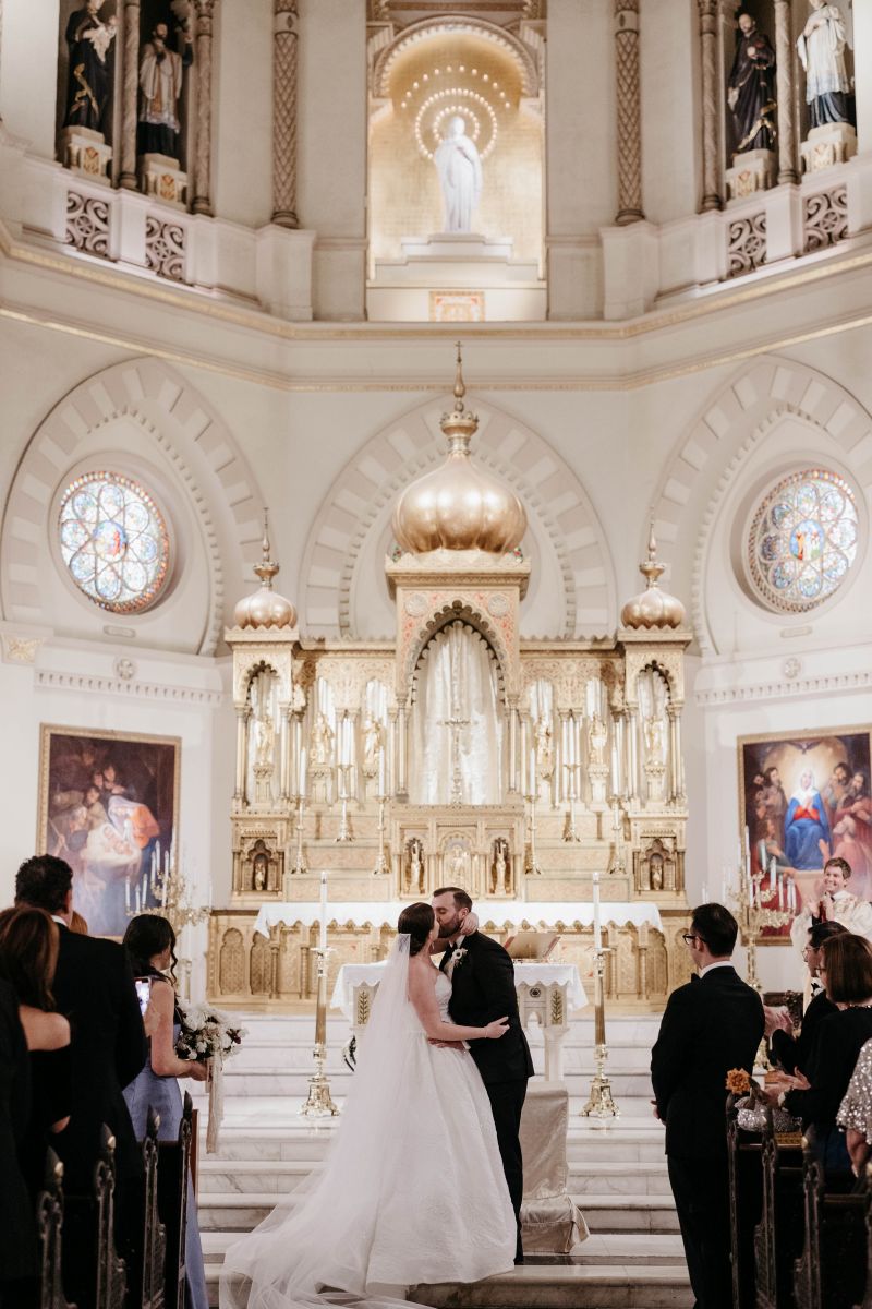 a couple kissing at their wedding ceremony in a church the woman is wearing a white wedding dress and veil the man is wearing a black suit
