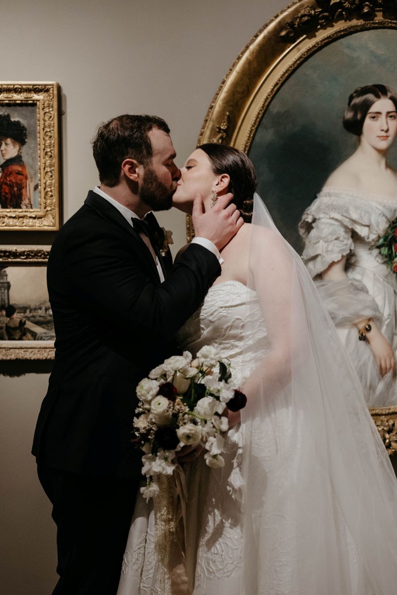 a couple kissing in front of art in a museum the woman is wearing a white wedding dress and veil and holding a bouquet of flowers the man is wearing a black suit
