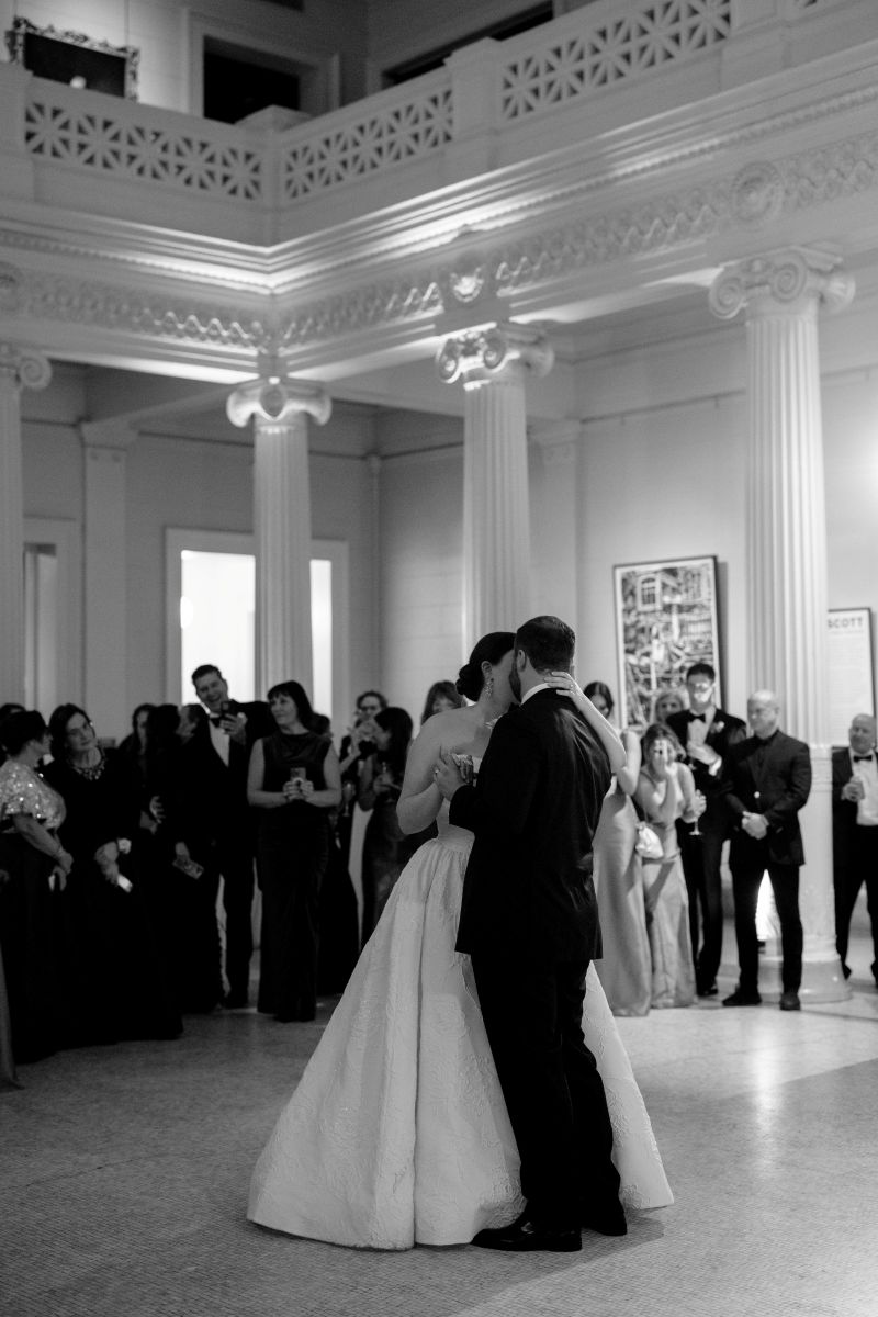 a couple dancing in a museum at their wedding reception the woman is wearing a white wedding dress the man is wearing a black suit as their guests watch them