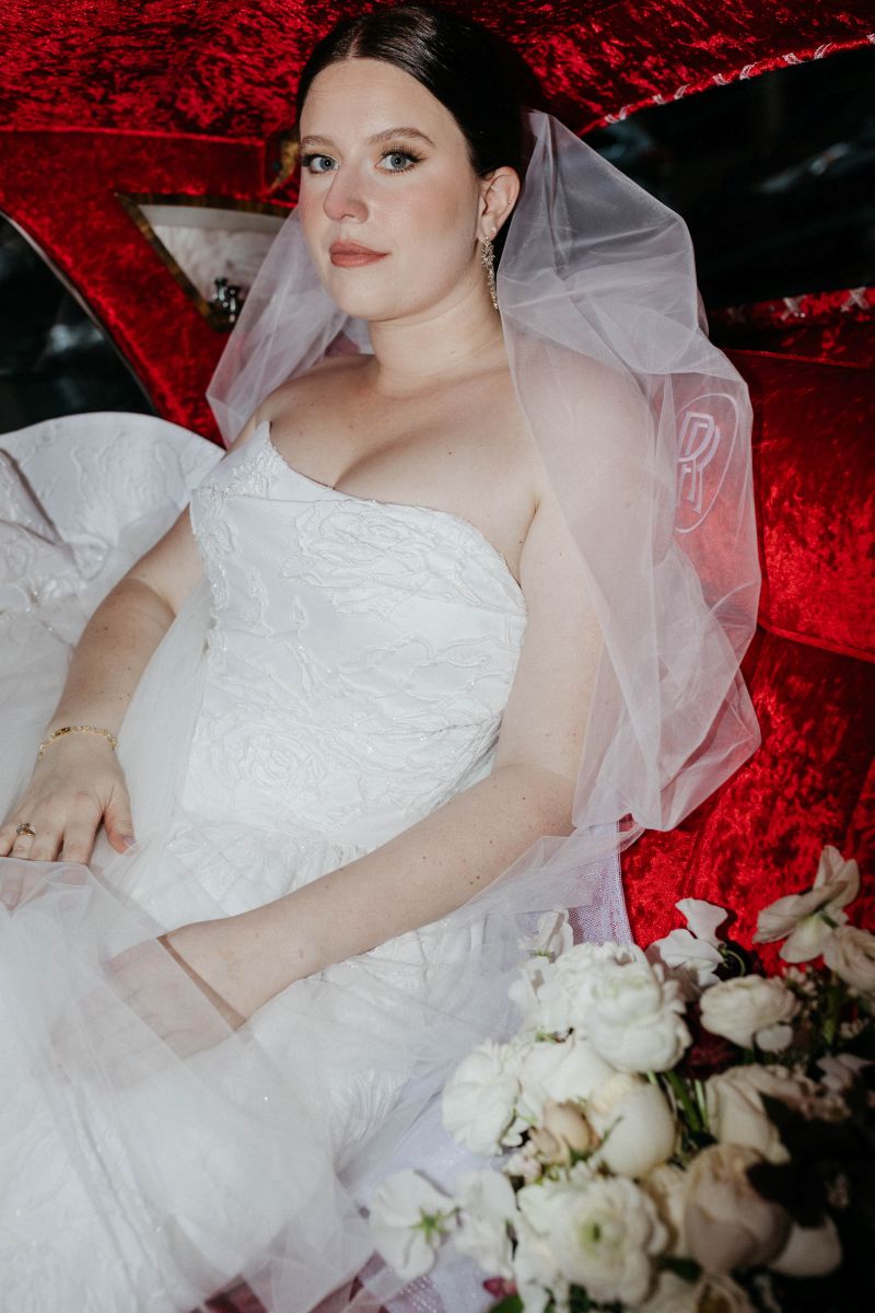 a woman inside a car softly smiling the car is lined with red velvet she is wearing a white wedding dress and veil