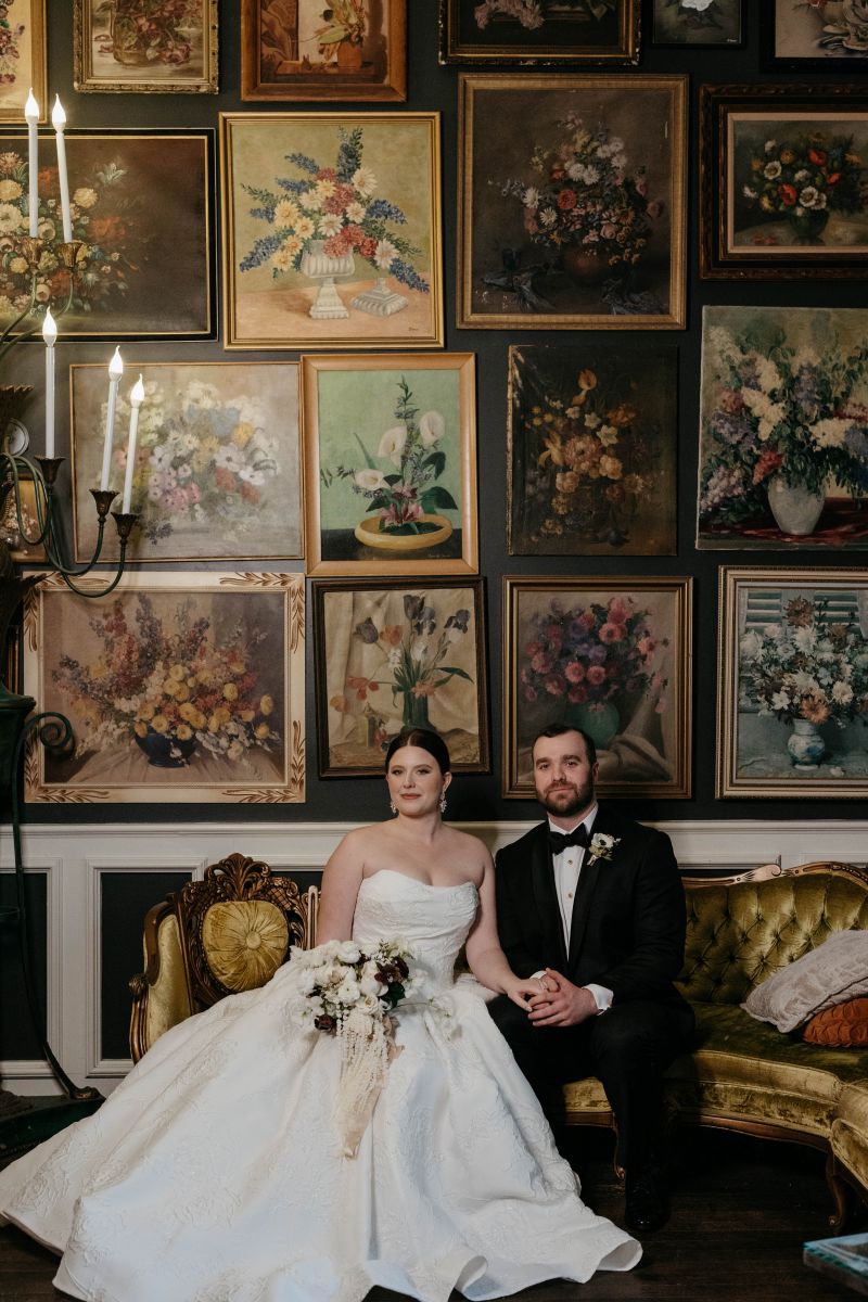 a couple sitting on a couch in front of a wall of art at the new orleans museum of art the woman is wearing a white wedding dress and holding a bouquet of flowers and the man is wearing a black suit
