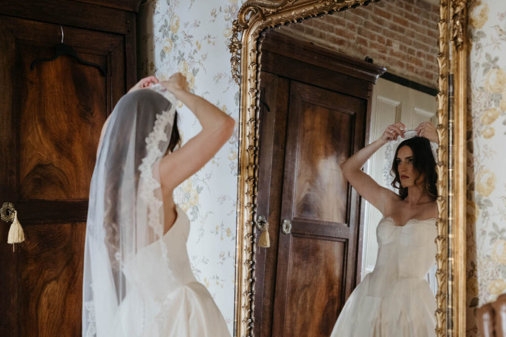 A person in a wedding dress looking into a mirror as they put on a veil