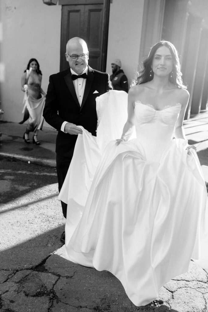 A person in a wedding dress walking across a street as someone holds up the back of their dress