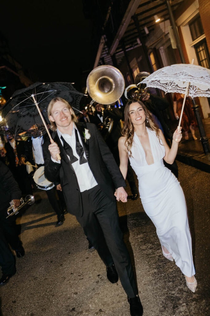 A newlywed couple holding hands and walking down the streets of New Orleans with a band behind them