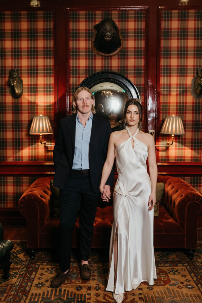 A couple holding hands and looking forward in front of a plaid colored wall
