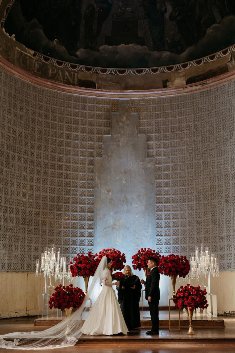 a couple having their wedding ceremony at hotel peter and paul in New Orleans at the altar with their officiant they are surrounded by red roses in gold vases and candles in clear candle holders 