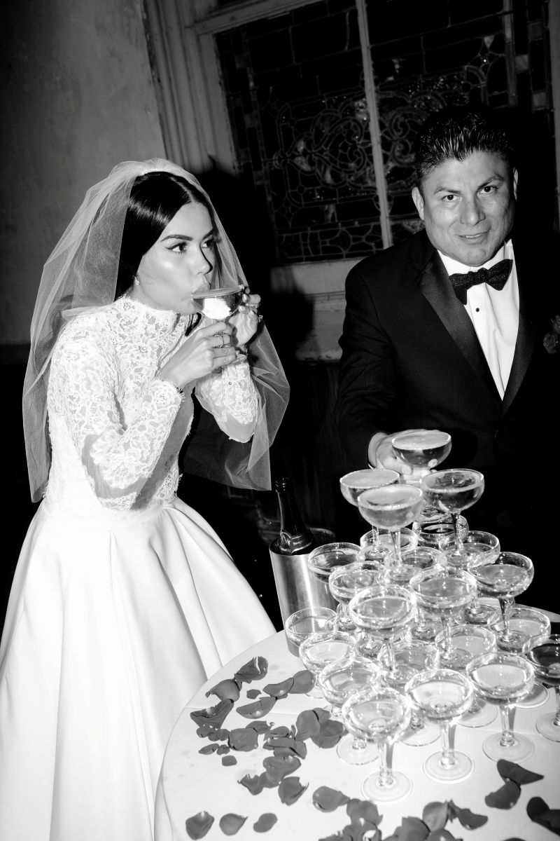 a bride drinking champagne from a champagne glass next to her partner in front of them is a table with a variety of champagne glasses 
