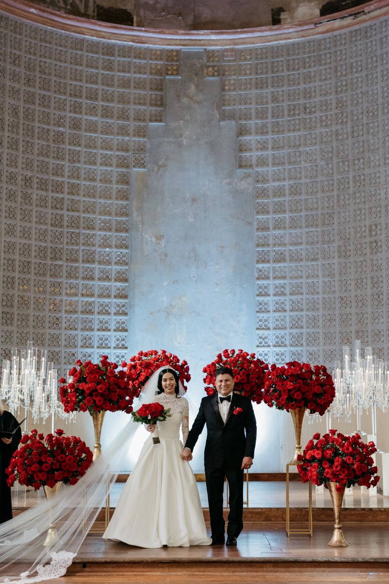 a couple holding hands at their wedding ceremony the bride is wearing a white wedding dress and veil and holding a bouquet of red roses her partner is wearin g a black suit they are both smiling and they are surrounded by red roses and candles 
