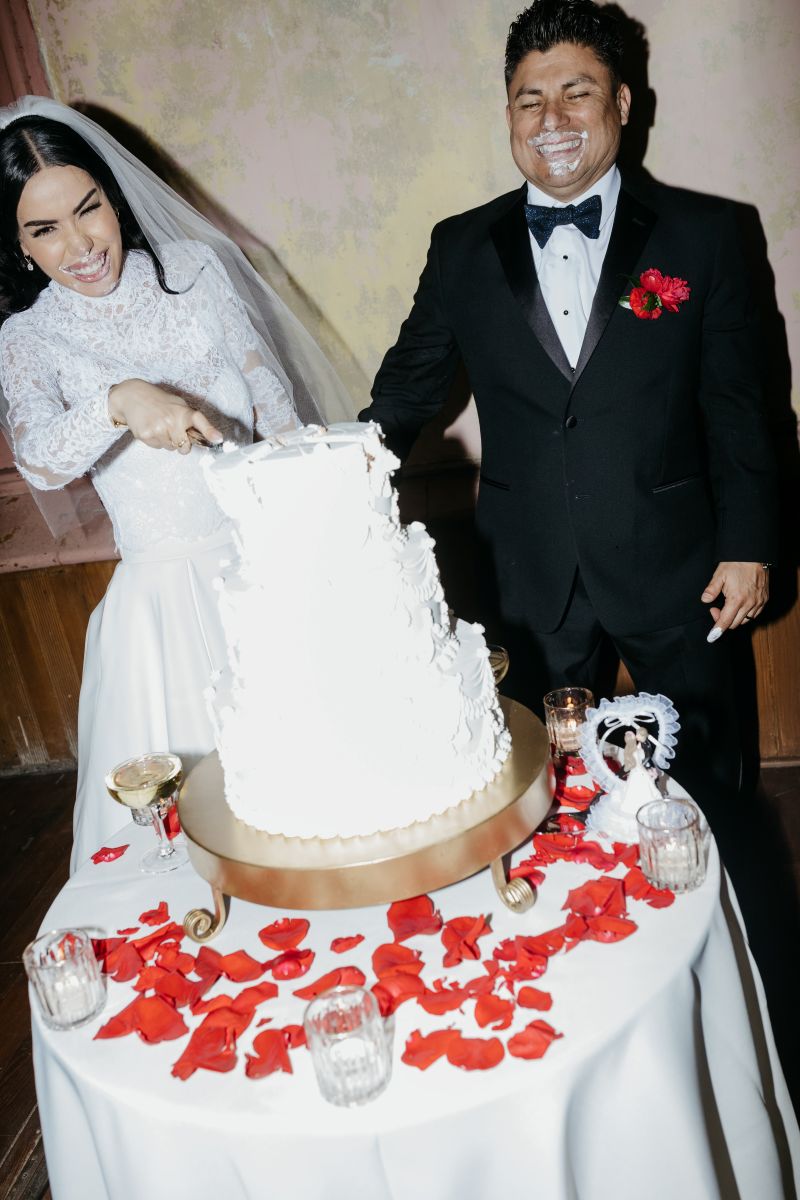 a couple enjoying their white wedding cake together the bride is cutting into the wedding cake while her partner laughs next to her