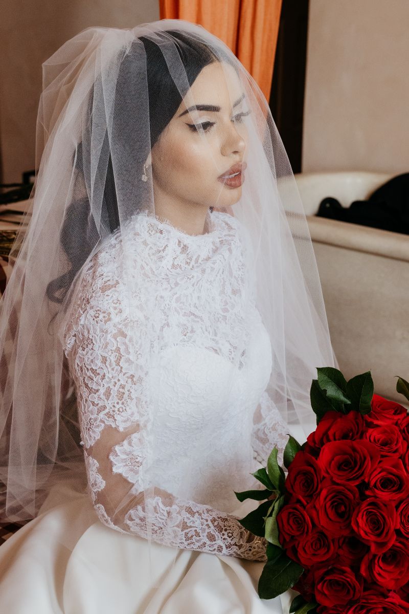 a woman sitting on a beige chair wearing a white wedding dress and veil and holding a bouquet of red roses in her hands 