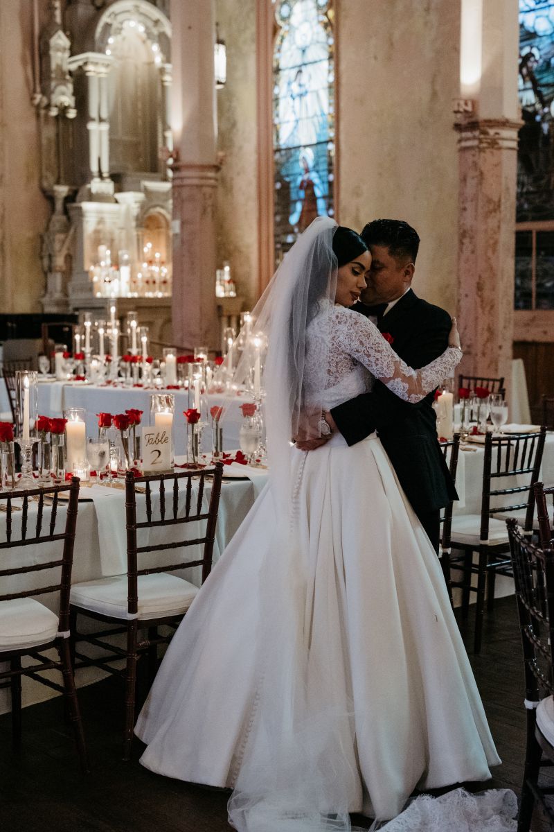a couple embracing inside of hotel peter and paul's chapel at their wedding reception next to banquet tables elegantly decorated with candles and roses the woman is wearing a white wedding dress and veil 