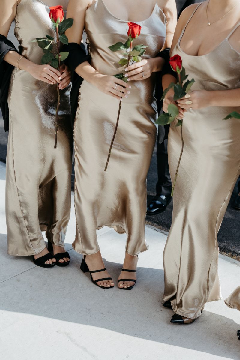 a trio of bridesmaids wearing gold dresses and holding red roses 