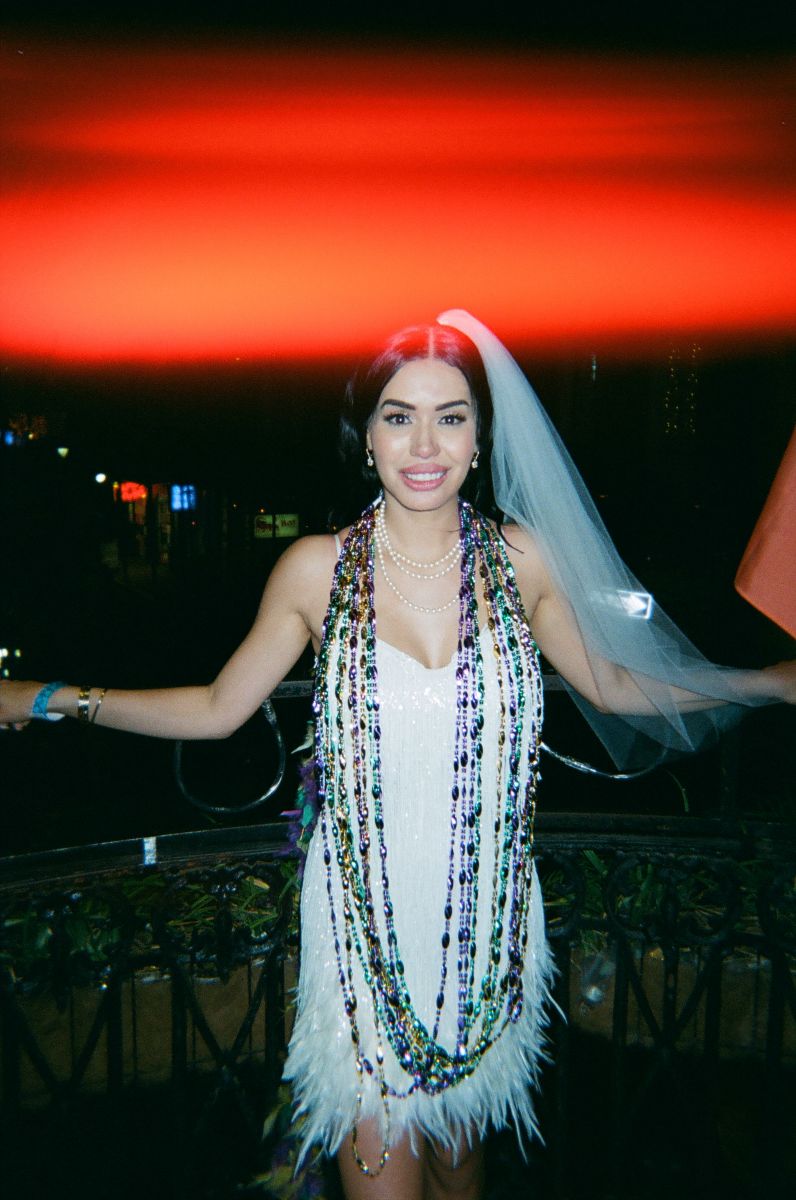 a woman wearing short white dress and veil and mardi gras beads standing on the balcony of cornet in new orleans 
