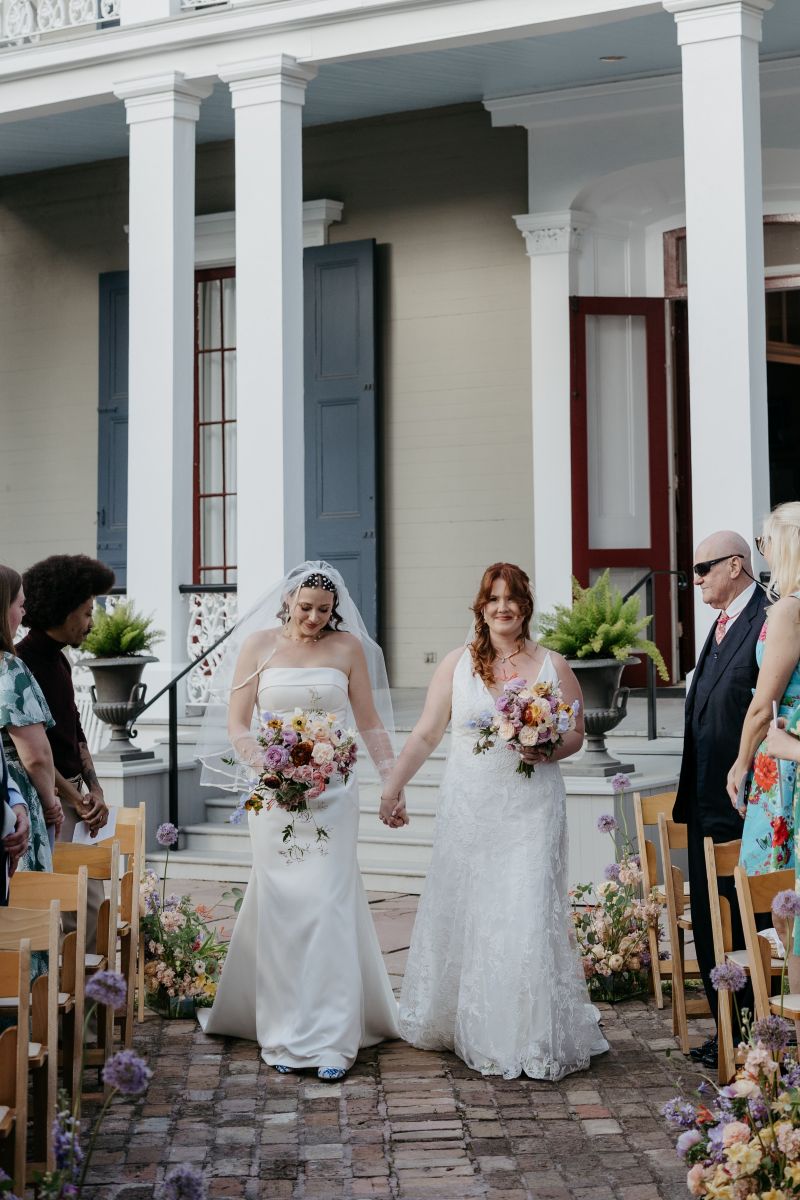 two women walking down the aisle together for their wedding at derbes mansion in new orleans they are walking hand in hand together in front of the mansion