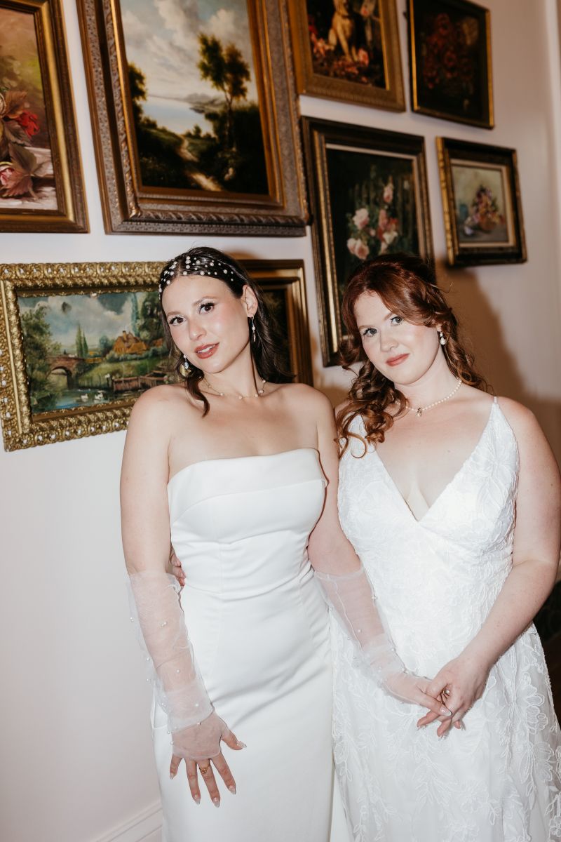 two women holding hands and wearing white wedding dresses standing in front of paintings in a hall