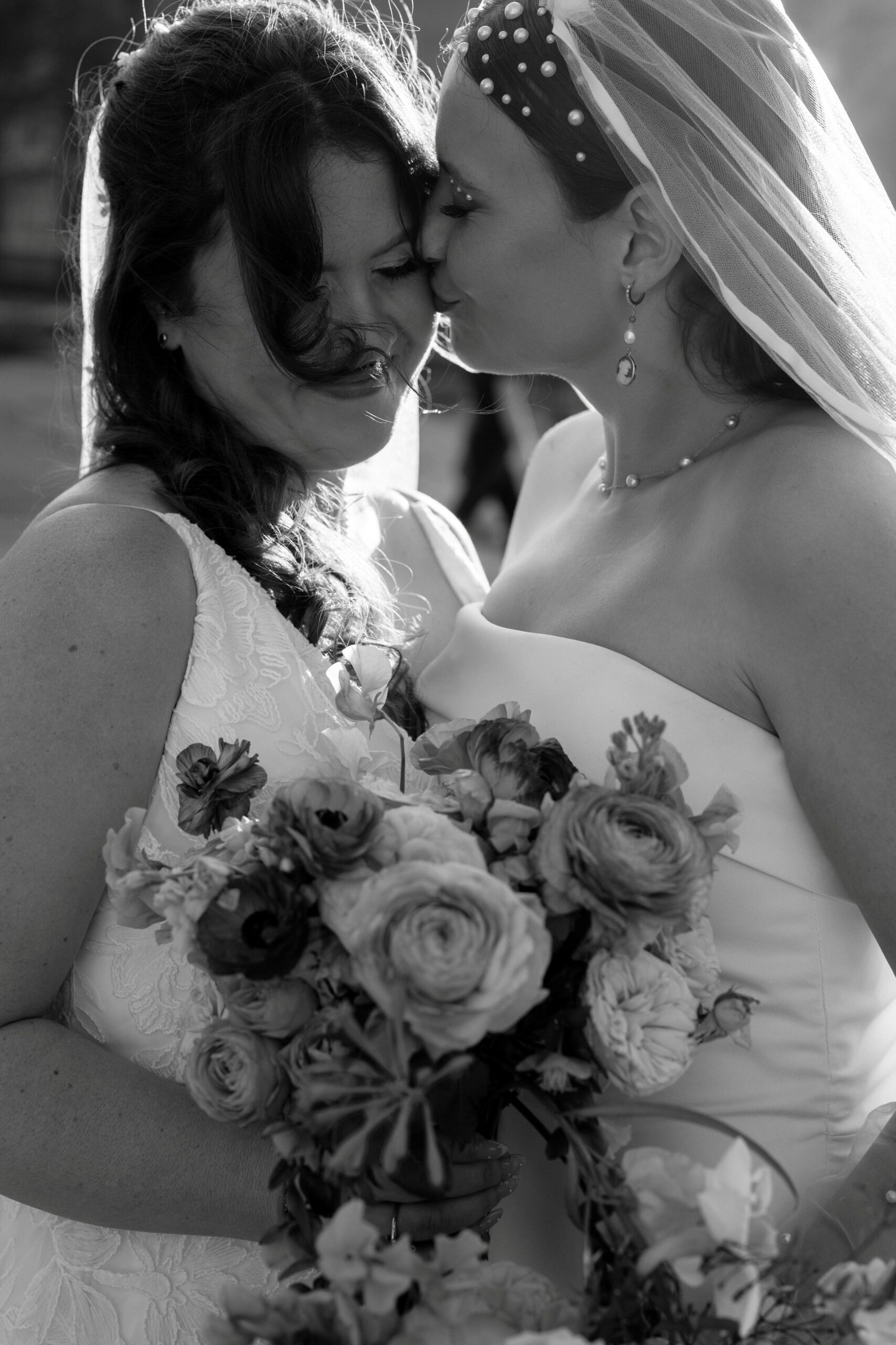 two women wearing white wedding dresses standing next to each other one is kissing the other on the cheek while they both hold a bouquet of flowers
