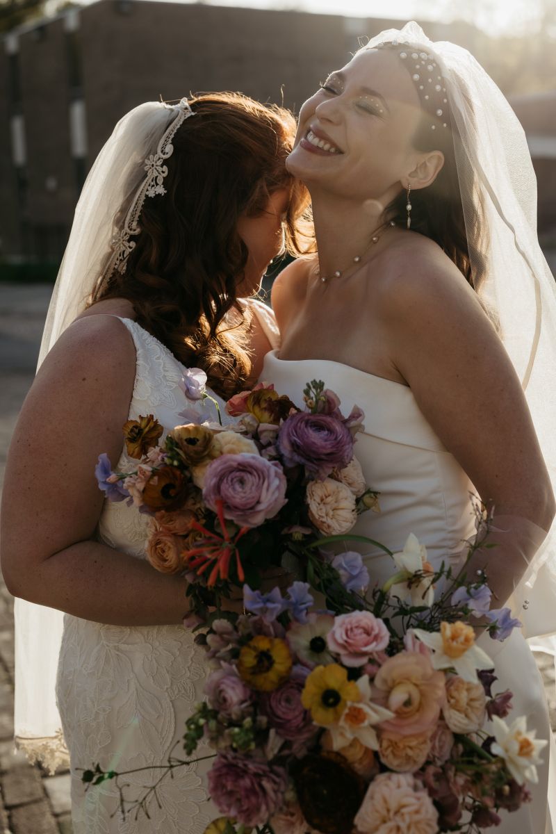 two women standing close to each other both wearing white wedding dresses and veils and holding flowers in their hands