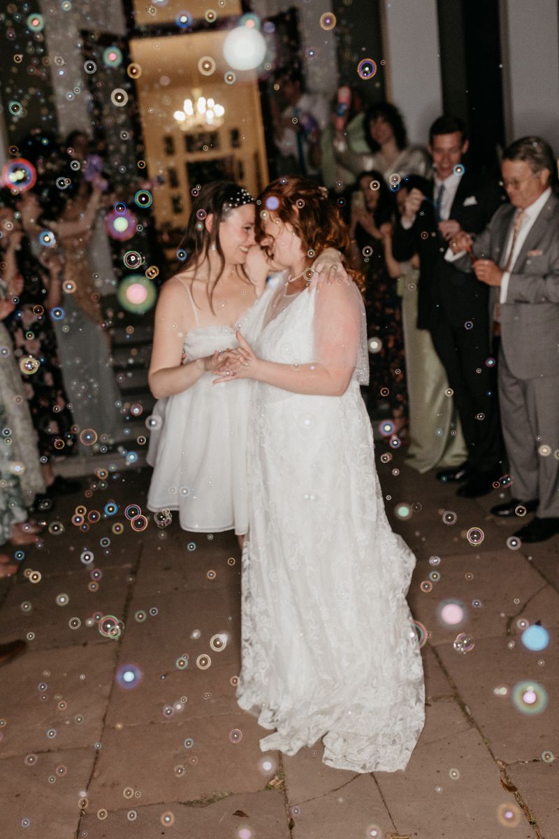 two women dancing together at their wedding reception while their guests blow bubbles and cheer for them