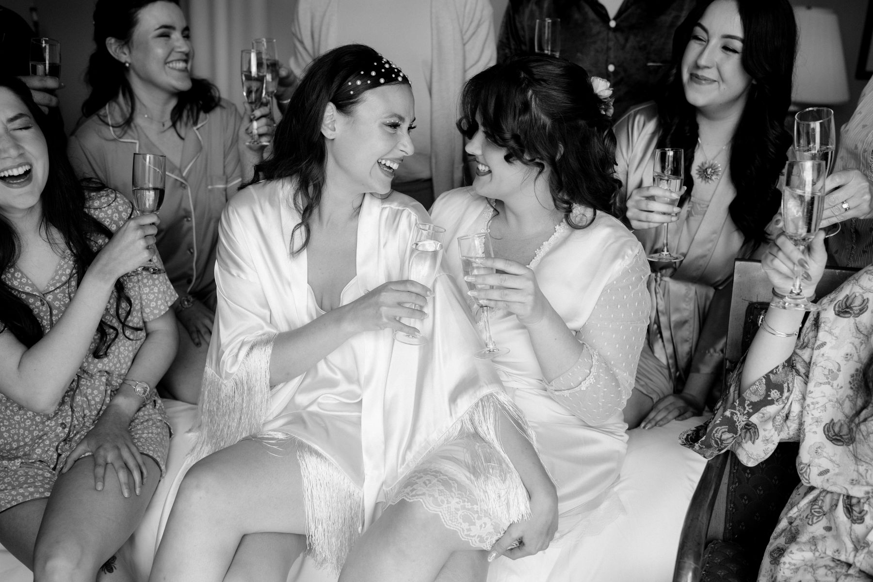 two women sitting on a couch together laughing and holding champagne glasses surrounded by their wedding party