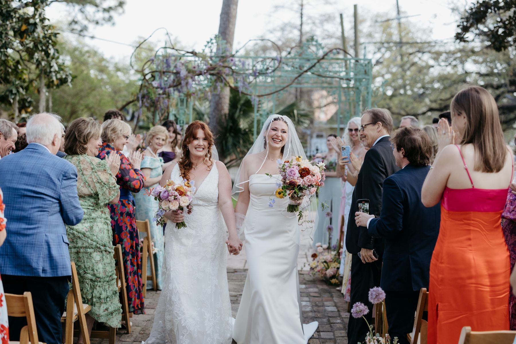 two women holding hands and smiling after their wedding ceremony at derbes mansion in new orleans as their guests stand and clap for them