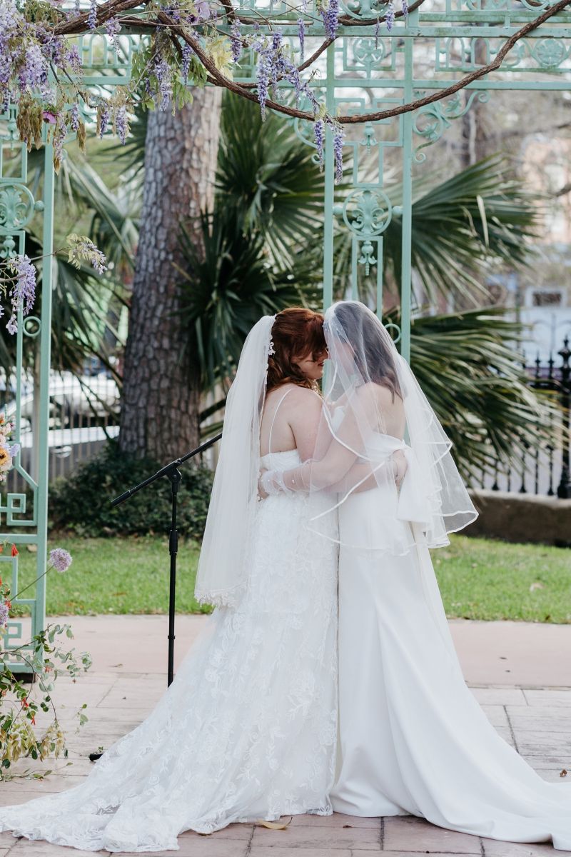 two women standing next to a wrought iron gazebo both wearing white wedding dresses and veils at derbes mansion in new orleans