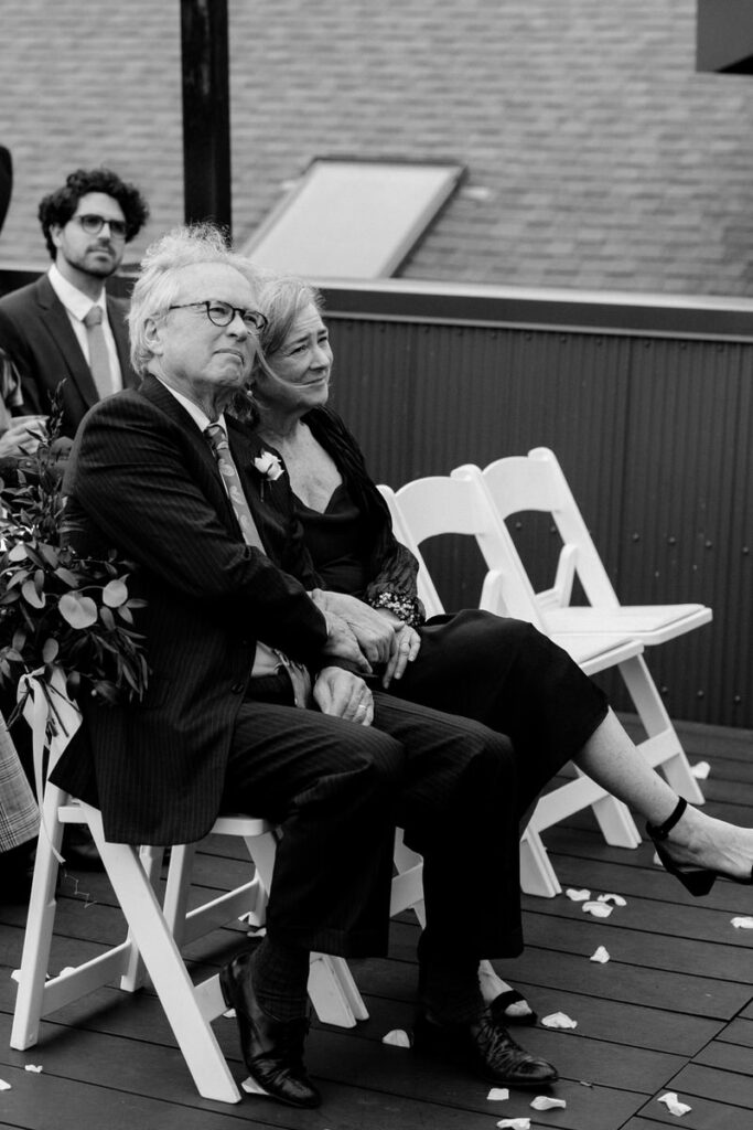 Two parents smiling as they watch a wedding ceremony 
