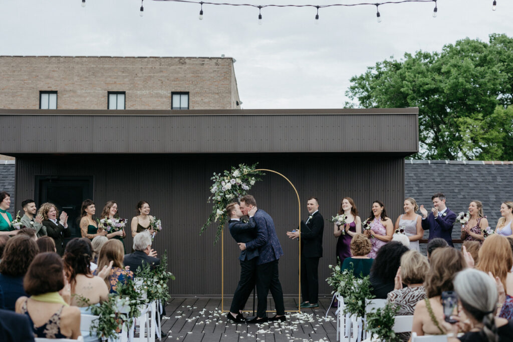 Newlyweds during their first kiss with their wedding guests and parties clapping 