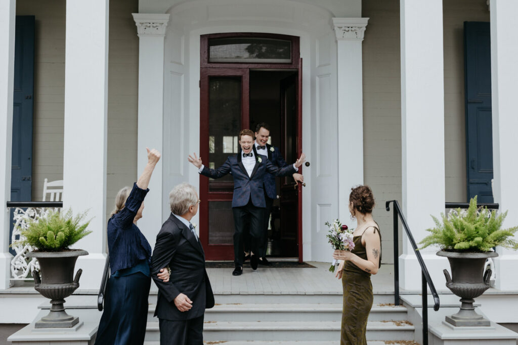 Two grooms walking out of a house with people cheering in front of them 