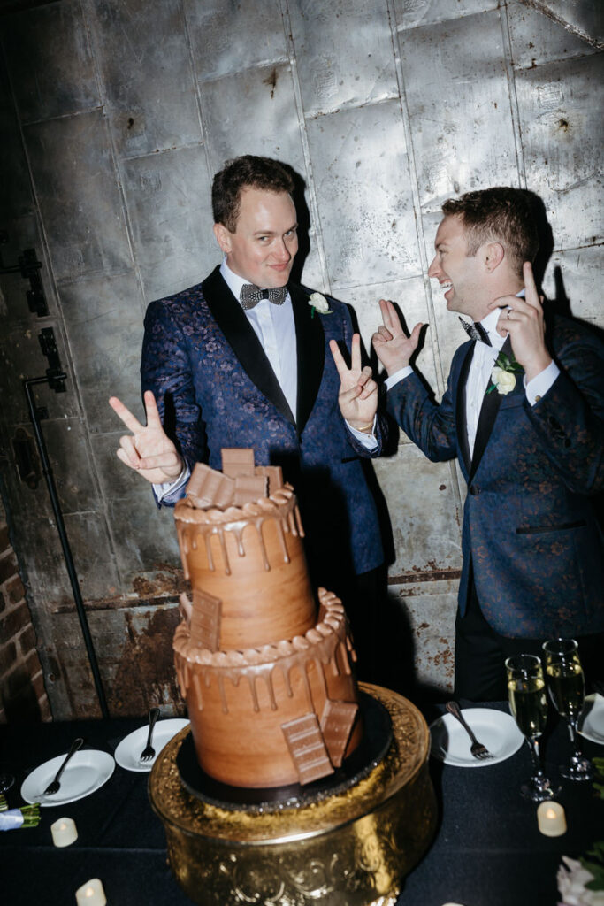 Newlyweds standing behind a two tiered wedding cake smiling and holding up peace signs 