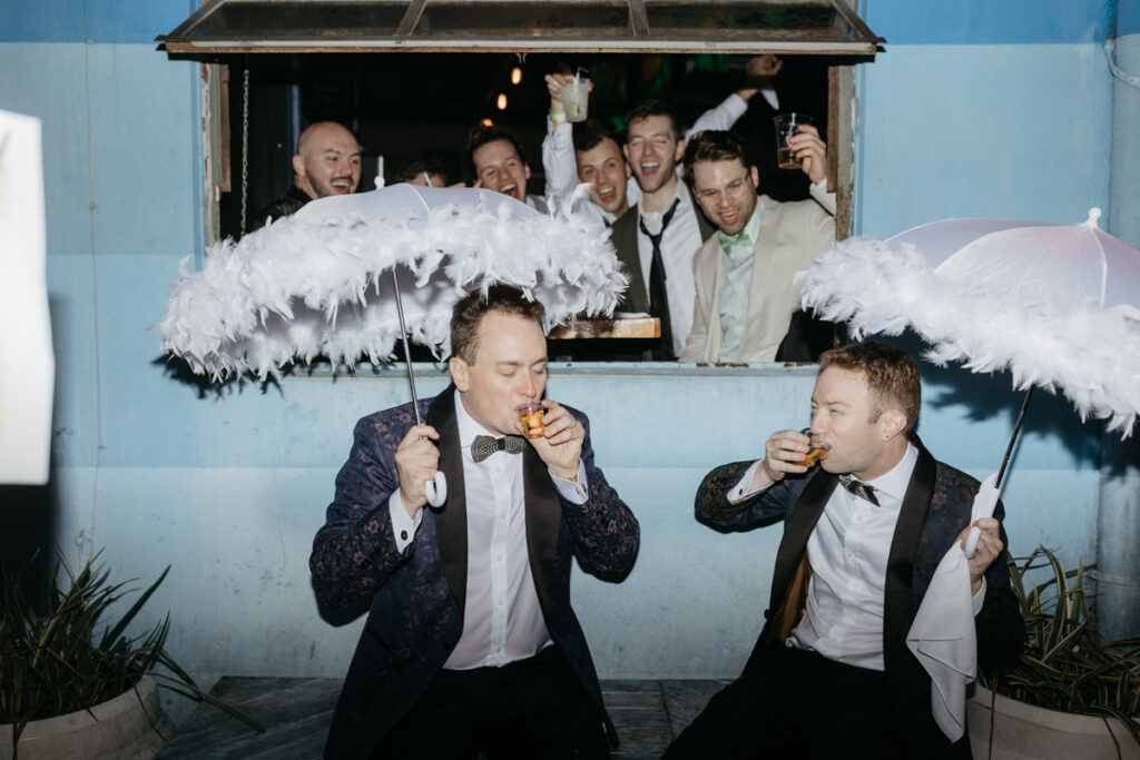 Newlyweds taking shots while their wedding guests cheer behind them 