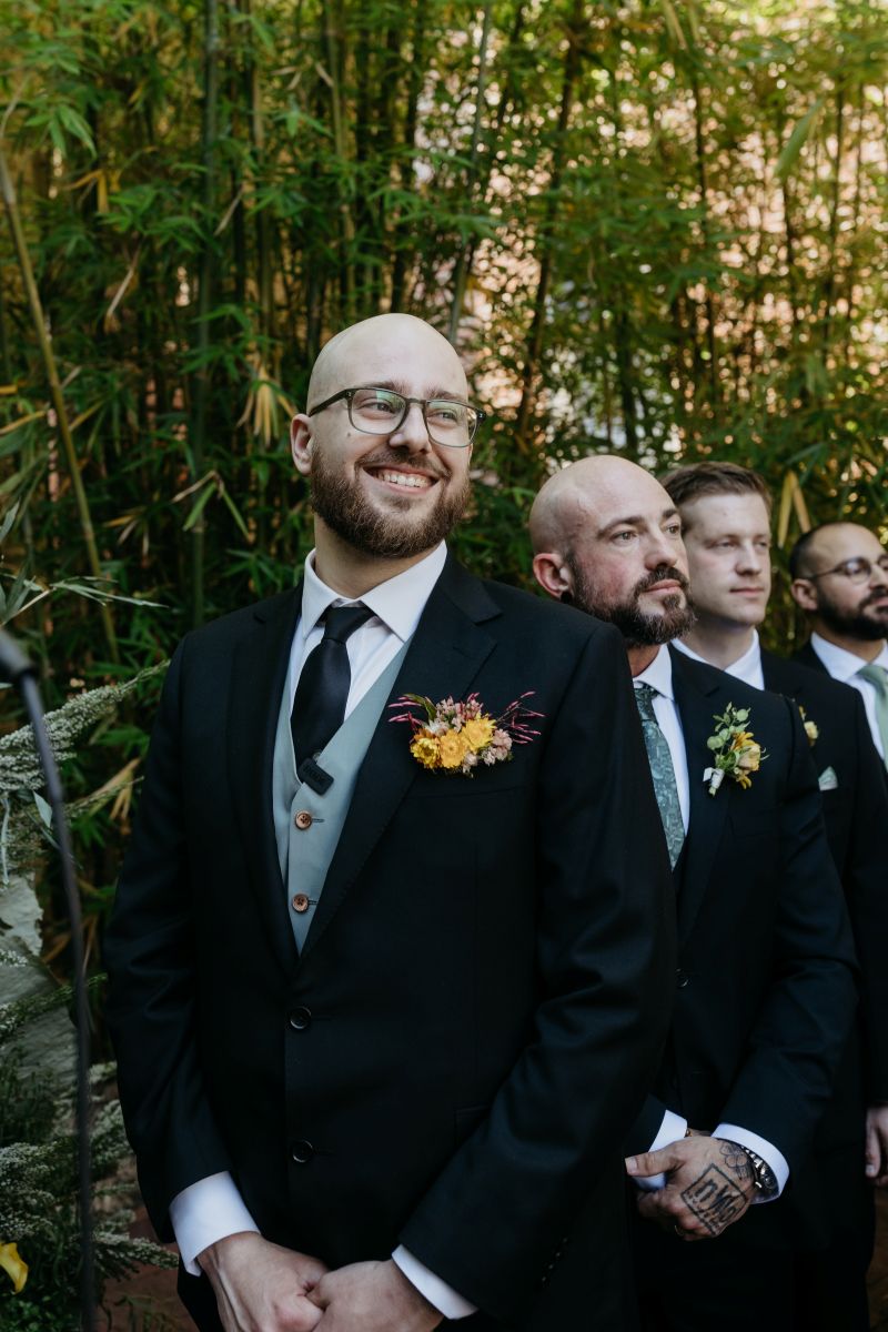 a man with his wedding party standing and smiling and waiting for his partner to walk down the aisle