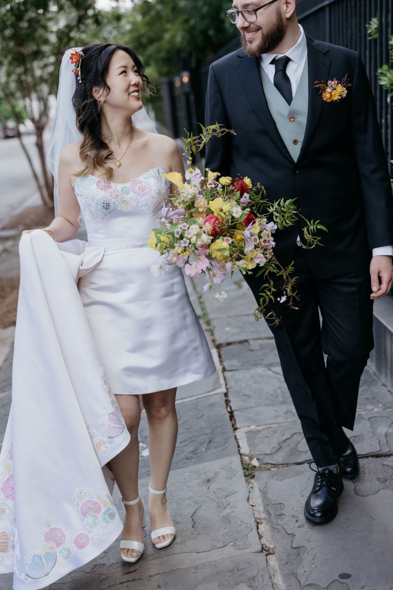 a couple walking down a street, smiling at one another the woman is wearing a white wedding dress and veil and holding a bouquet of flowers the man is wearing a suit