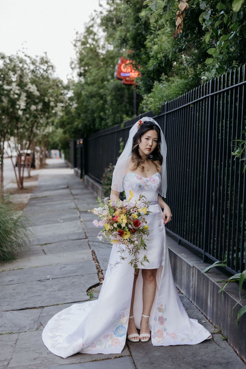 a bride wearing a white dress and veil standing on the sidewalk and holding a colorful floral arrangement