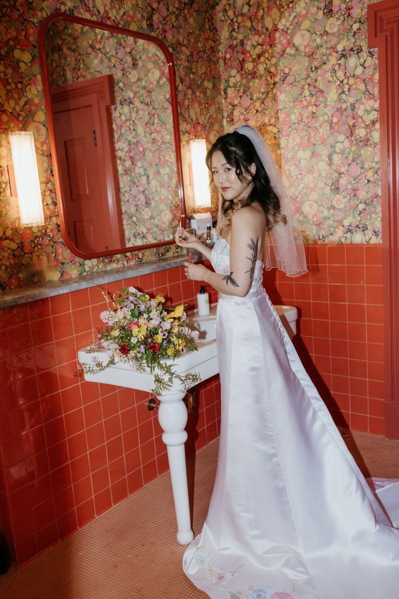 a woman in a bathroom in hotel saint vincent in new orleans getting ready for her wedding and wearing a white wedding dress and veil her wedding bouquet is resting in the sink