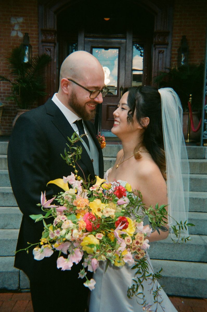 a couple standing in front of the steps at hotel saint vincent on the day of their wedding they are smiling and laughing with each other and the woman is holding a colorful floral bouquet